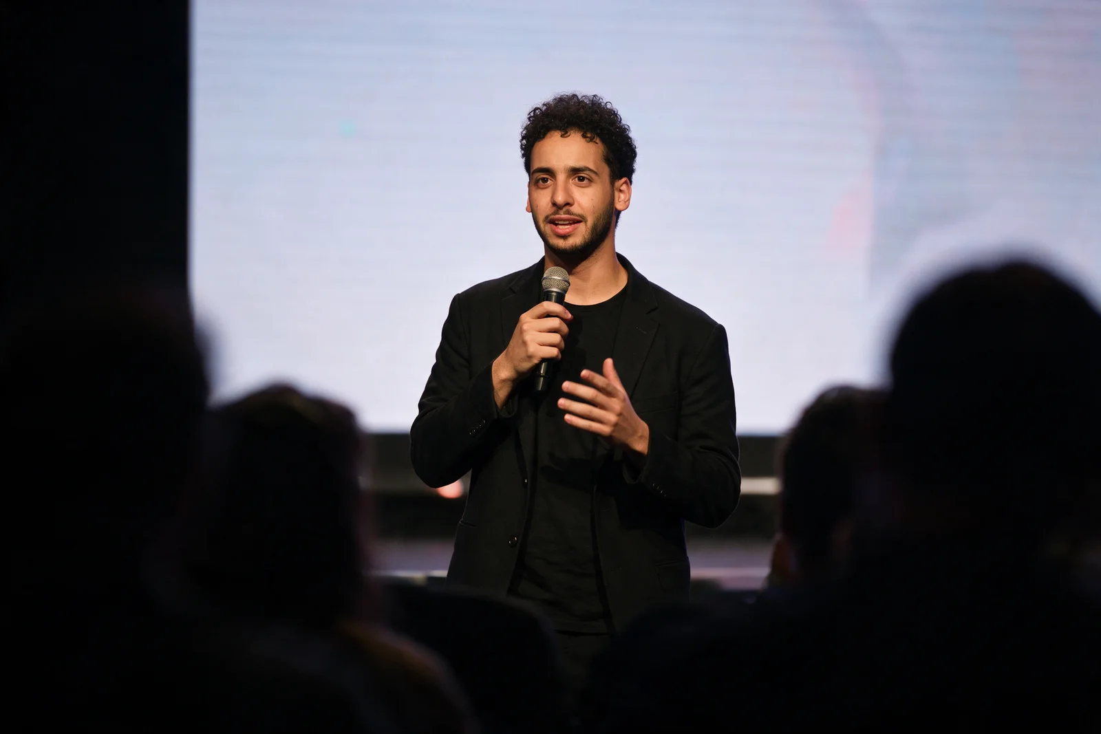 Zein Kaskas speaking on a tech-conference stage with a handheld microphone, a glowing screen behind him and audience silhouettes in the foreground.