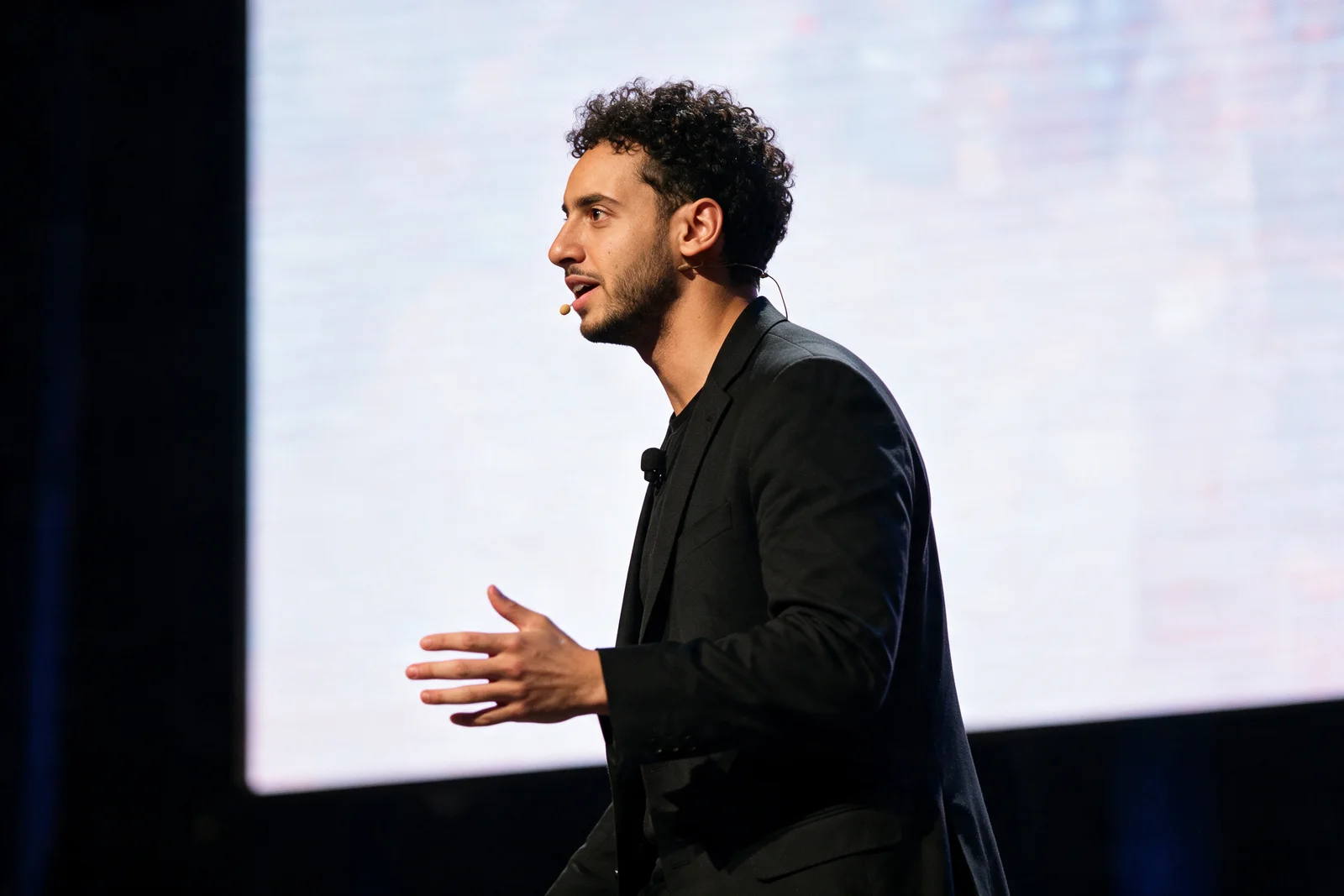 Zein Kaskas walking a conference stage in profile while speaking into a lav mic, a large glowing screen behind him.