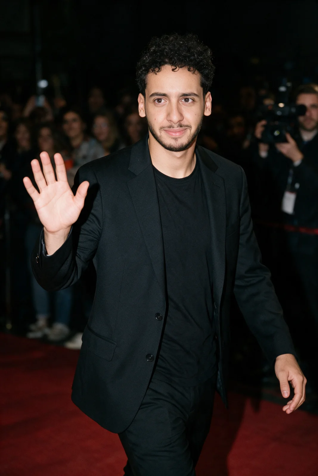 Zein Kaskas waving as he walks a red carpet at night under flash photography.