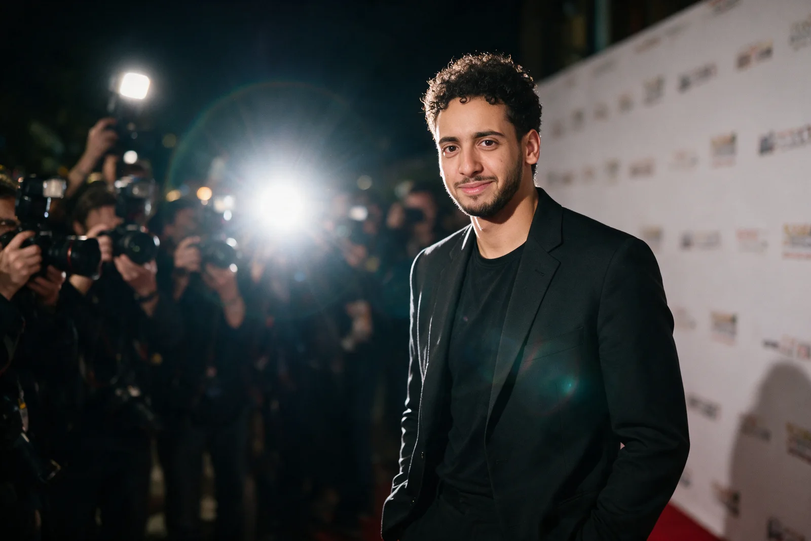 Zein Kaskas pausing on a red carpet with his hands in his pockets, turned toward a wall of photographers.