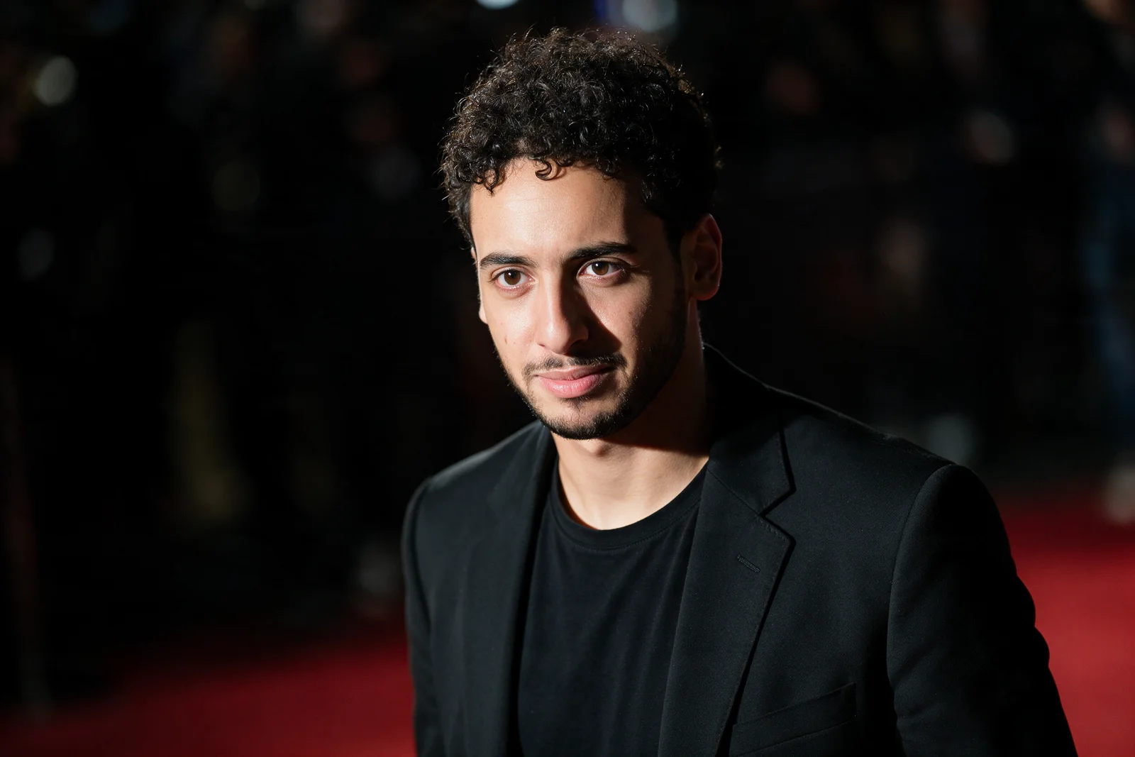 Tight waist-up shot of Zein Kaskas on a red carpet at night, a bright flash catching one side of his face.
